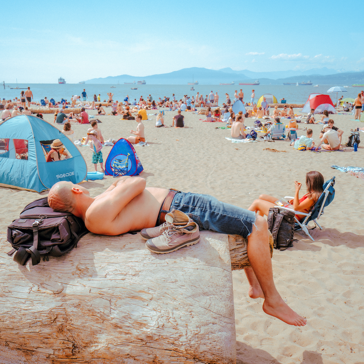 A man lies on a log at the beach on a busy day. The water and the mountains are visible in the background. It is very sunny.