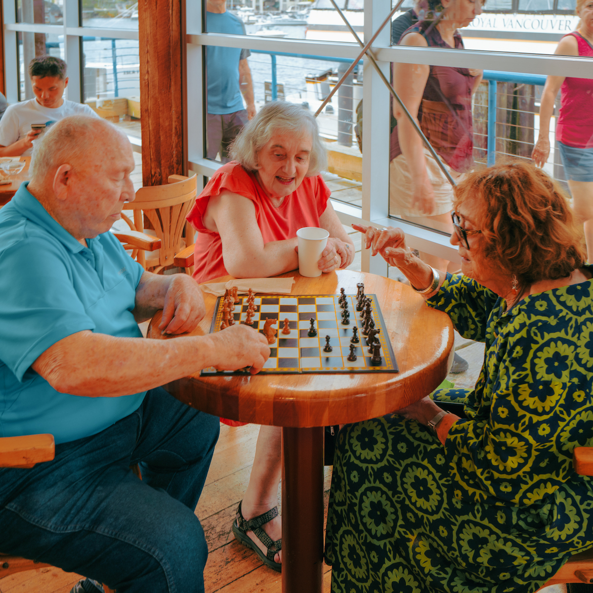 Two seniors play chess at a food court while a third watches.