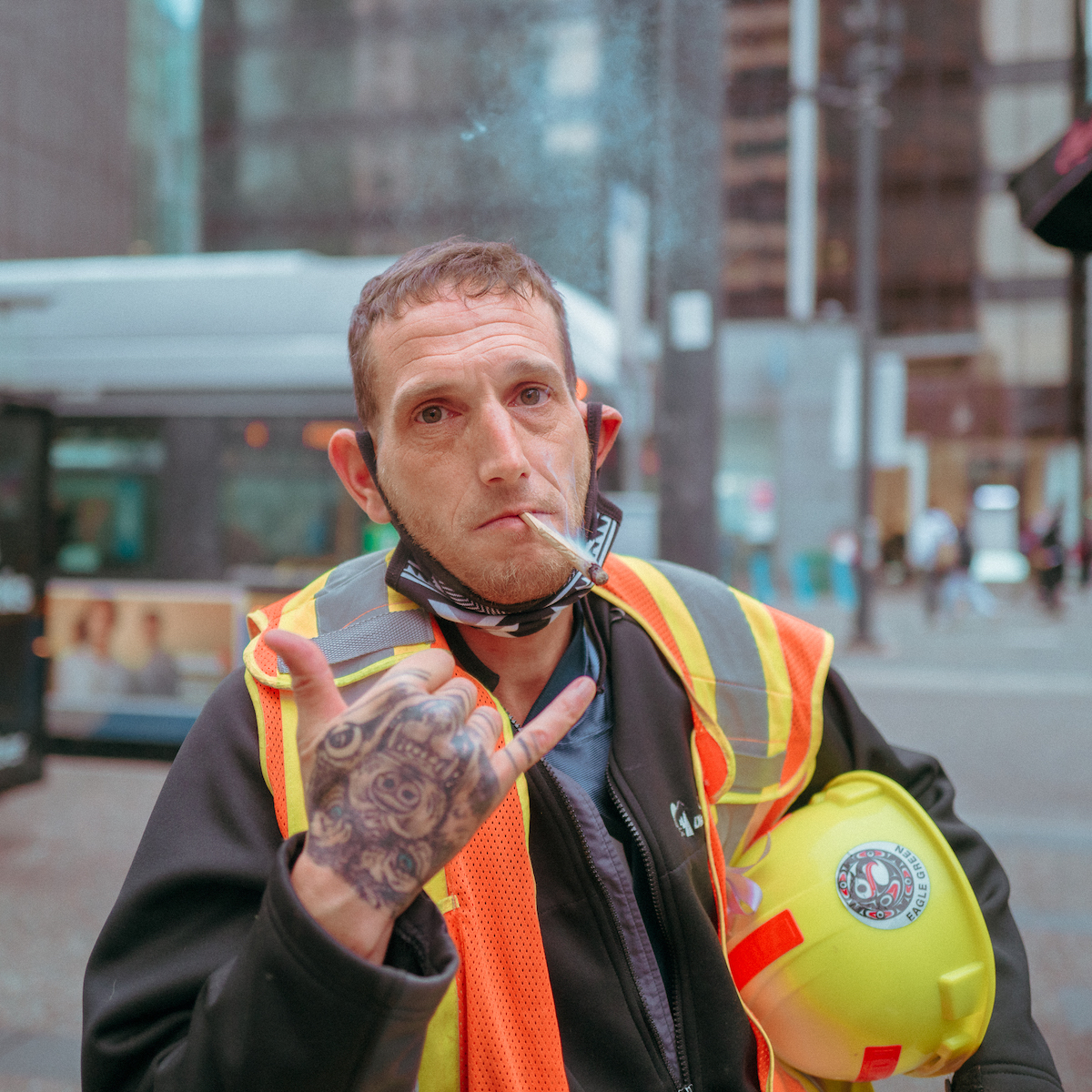 A construction worker in an orange vest holding his yellow helmet pauses to do the “hang 10” sign to the camera. The back of his hand is covered in tattoos.