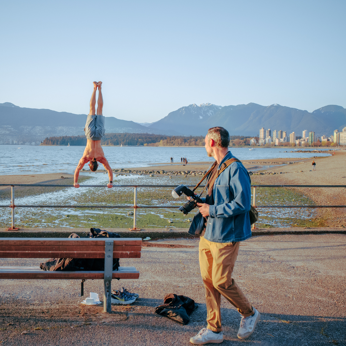 A photographer at the beach walks past a man doing a handstand on a fence.