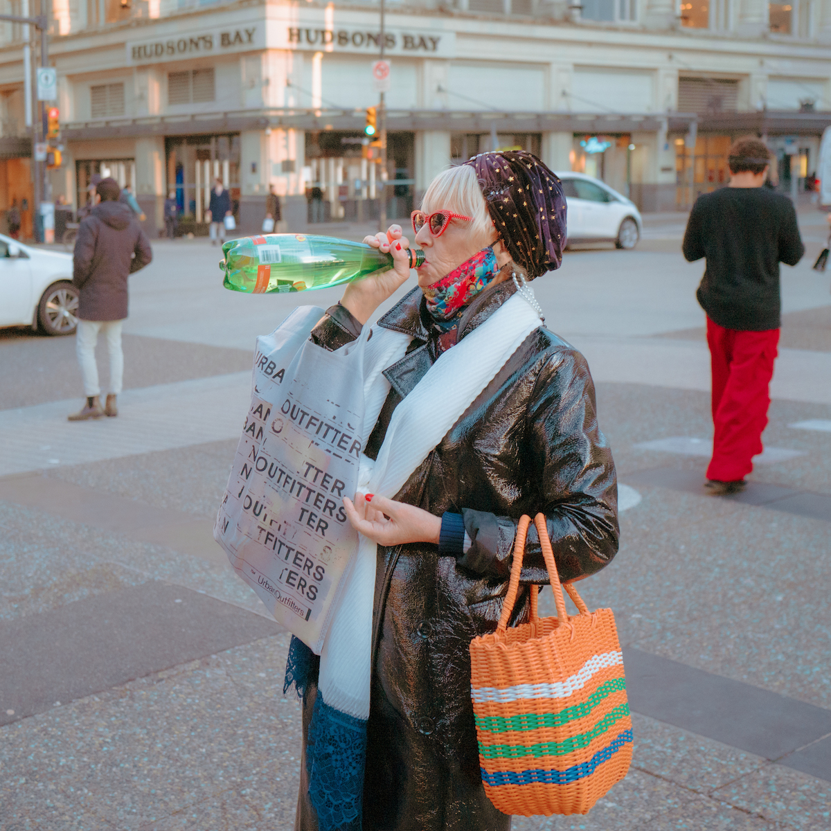 A woman in a big leather coat and red sunglasses pauses at an intersection to guzzle Perrier.