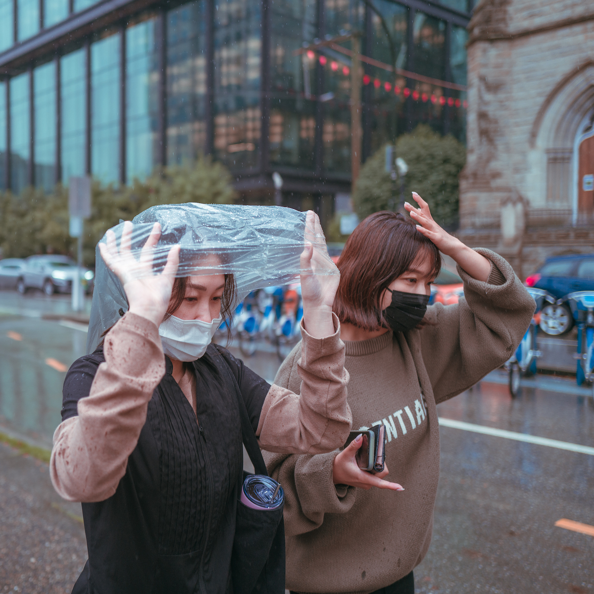 Two young women are caught in the rain. One uses her hand to shield herself. The other uses a plastic bag.