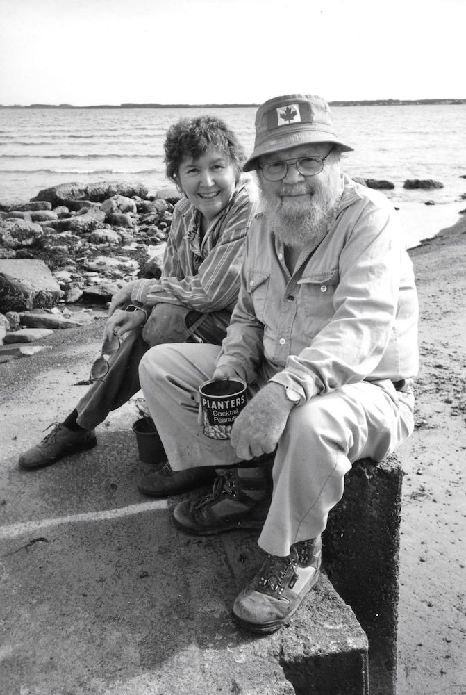 A black and white photo shows an older Claire and Farley Mowat sitting on a low structure by the beachside.