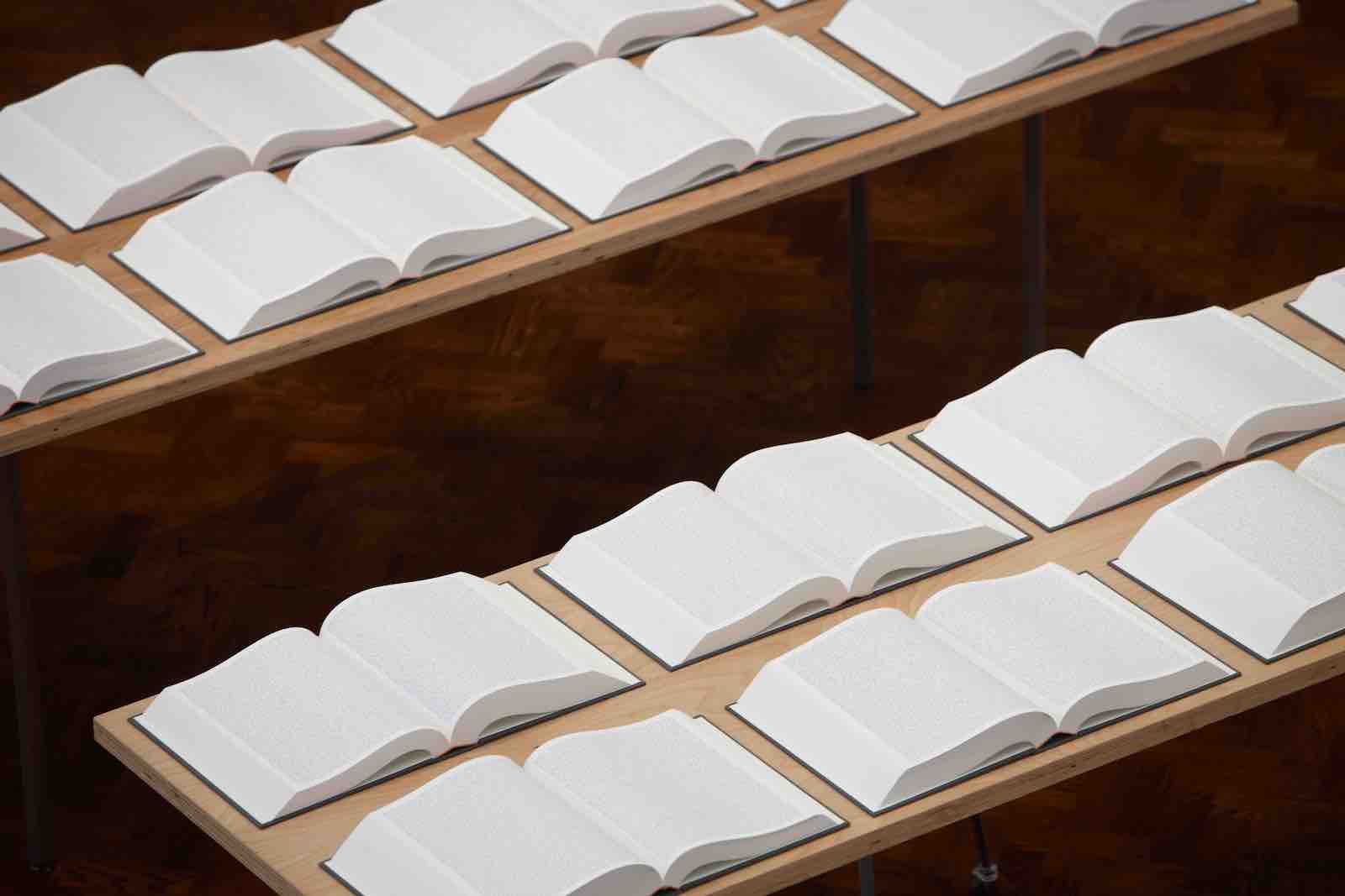 Numerous books with small text on white pages lie open in neat rows on two long wood tables.