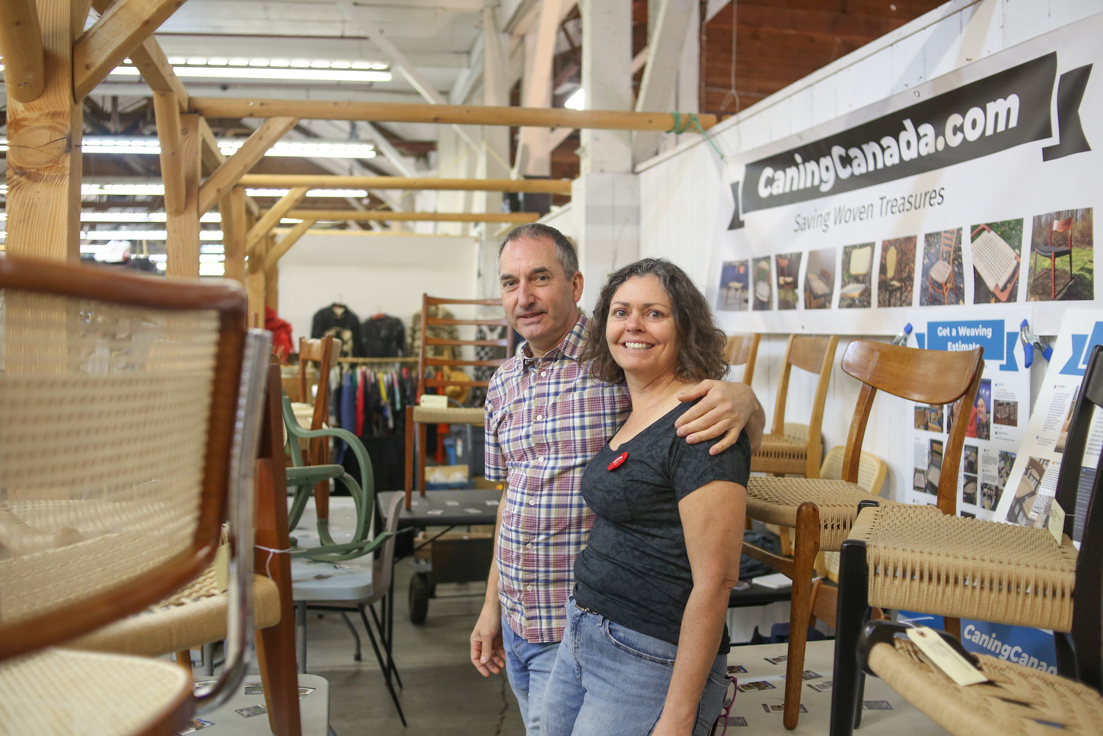 A man with his arm around a woman inside an old barn that has been converted into a flea market. Around them on tables are corded chairs.
