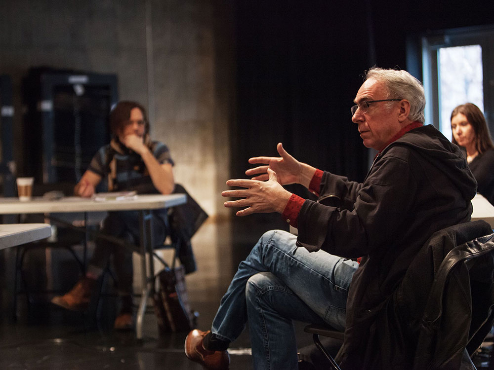 Norman Armour sits in a chair to the right of the frame, and his face is visible from the side. He has white hair and glasses and is wearing a black hoodie over a red button-down shirt, his arms outstretched in conversation. In the background are two people seated at white tables, listening. The studio space they are in is dimly lit with dark concrete walls.