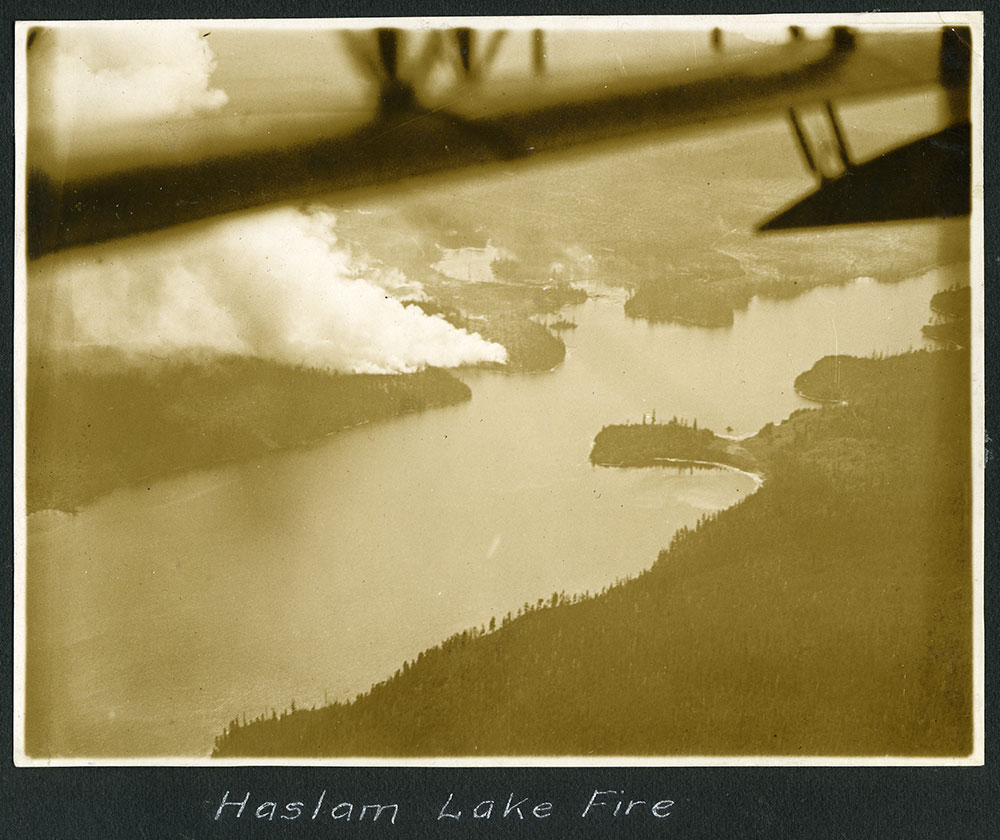 A sepia-toned photo reads 'Haslam Lake Fire.' The photo has been taken from a float plane &mdash; the wing is visible &mdash; as trees ringing Haslam Lake send up plumes of smoke.