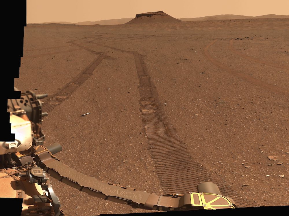 A panoramic view of a flat red sandy surface against a light orange sky. The left side of the frame is covered in black pixels. In the foreground to the left of the frame is steel equipment on the exterior of a surveying vessel.