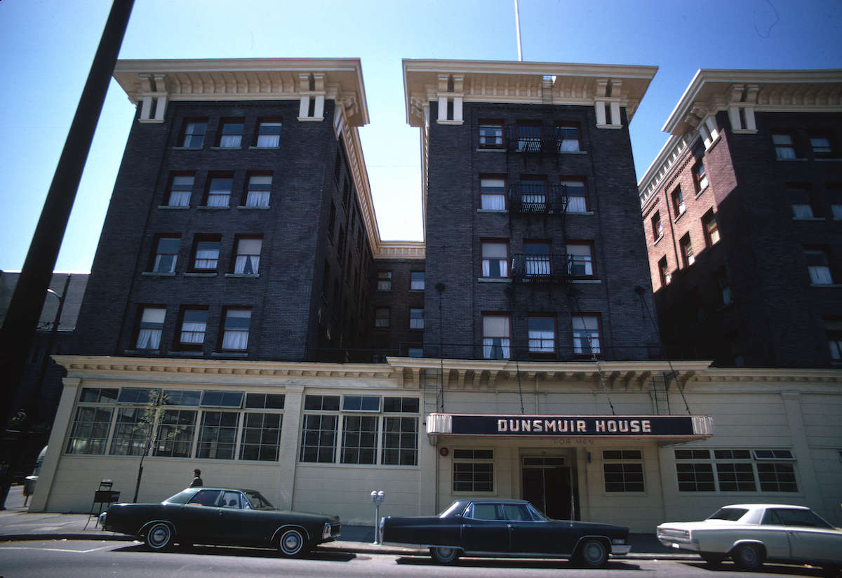 A photo of the front of the Dunsmuir. It is in colour but has a bit of a sepia/brownish '70s vibe. Cars from the era are parked in front of the building, which now bears the name and sign 'Dunsmuir House.'