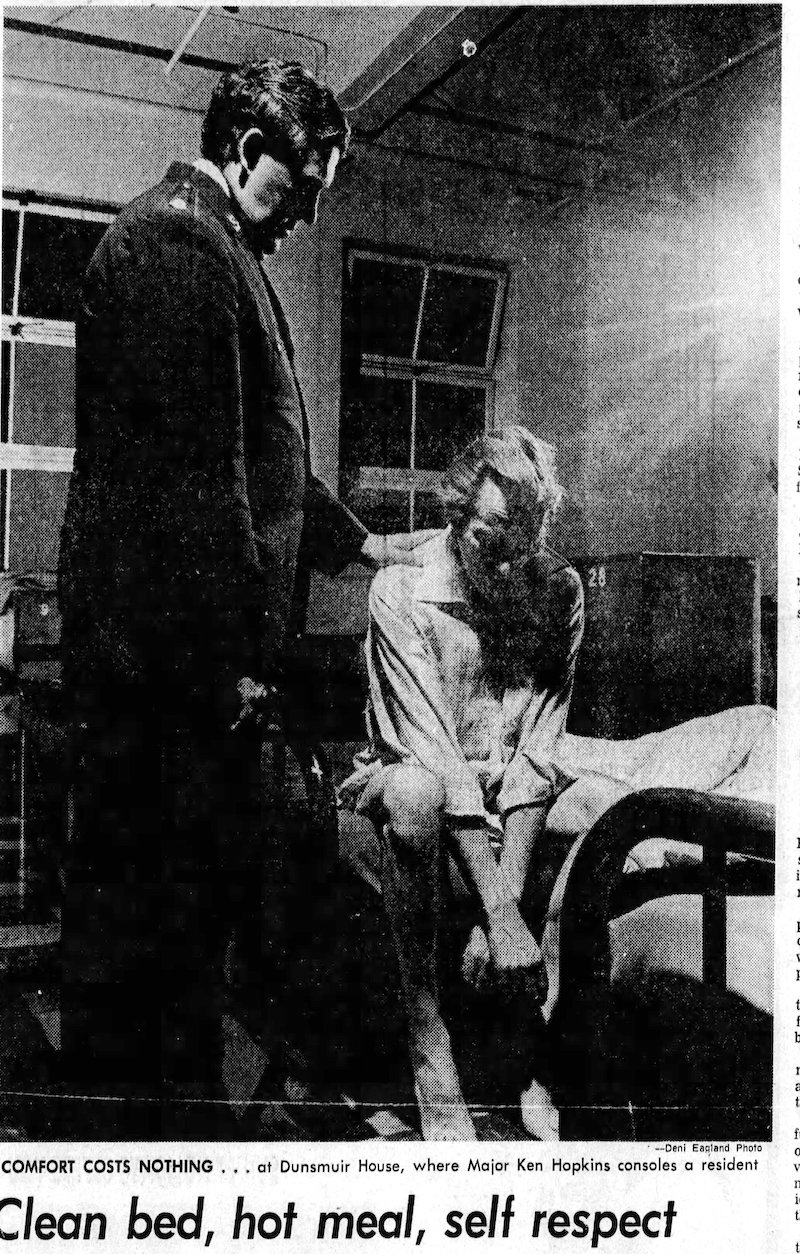 A black and white photo shows a man sitting on a bed, hunched over. Another man, dressed in a suit, stands beside him with his hand on his shoulder.