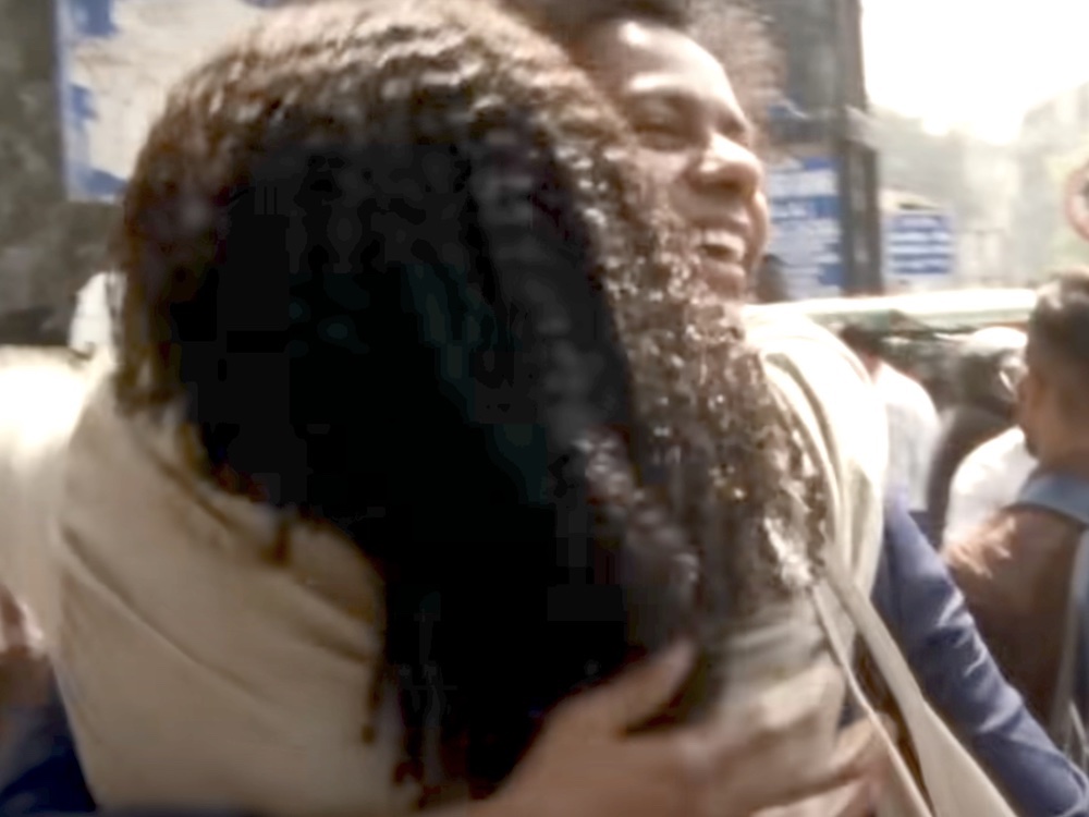 Two Black people embrace each other on a busy downtown street on a sunny day, with one person&rsquo;s back turned to the camera. The person who is facing the camera is looking up, smiling brightly.
