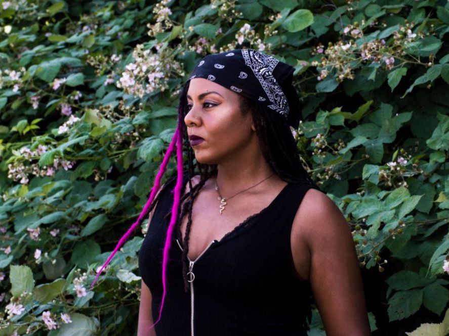 Ruby Smith D&iacute;az is a Black woman with long black hair and bright fuchsia highlights. She wears a navy kerchief in her hair and a black sleeveless top. She stands against a sprawling green blackberry bush with white blossoms. She looks serenely towards the left of the frame.