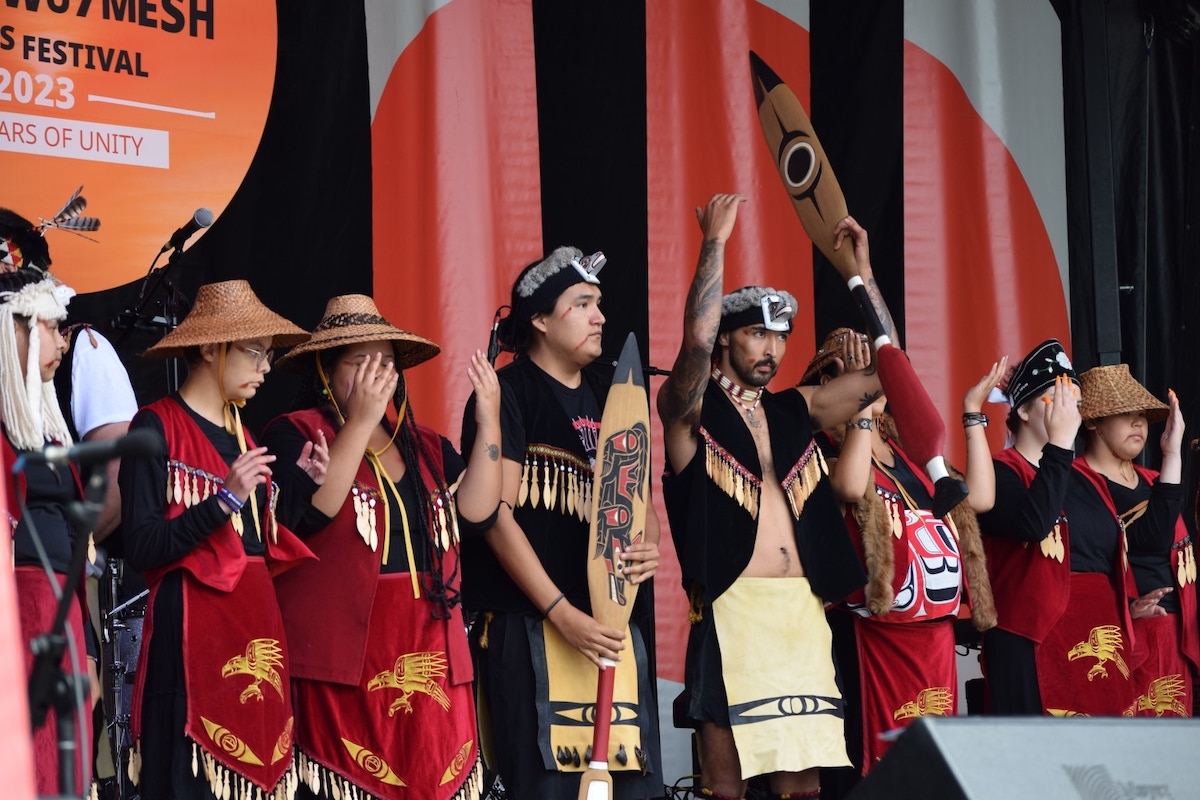 People onstage wearing Sḵwx̱wú7mesh clothing hold their hands up.