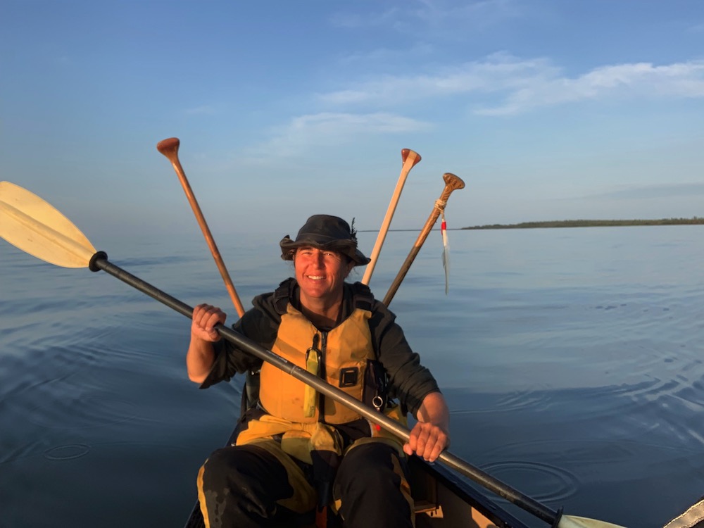A woman in a yellow lifejacket, black hat and black top paddles towards the camera in a canoe. Behind her is an expansive blue sky and a wide stretch of water on a sunny day.