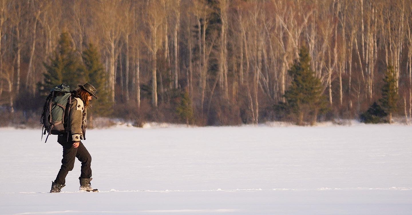 A woman to the left of the frame wears a brown wool jacket, hat, backpack and hiking boots. She steps across a stretch of snow against a stand of trees in golden light.