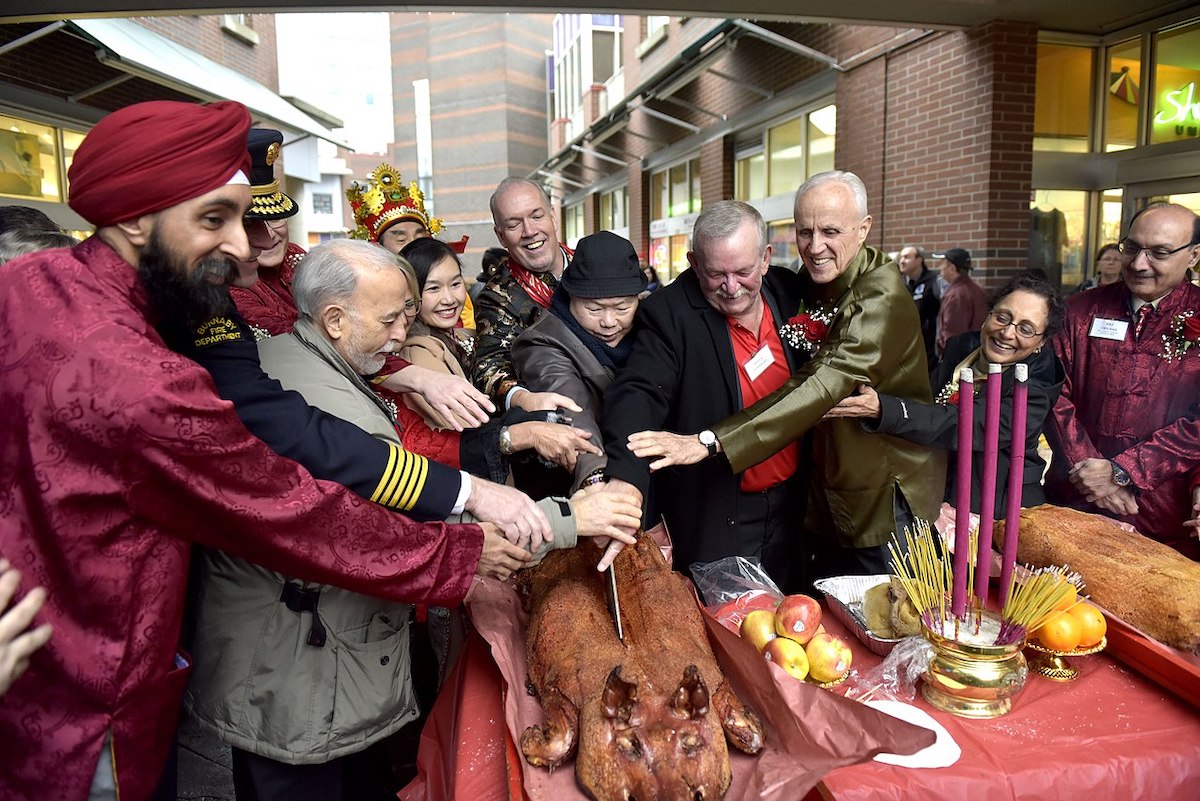 Ten or 12 people gather for a photo op that captures making the first slice into a roasted pig. John Horgan is at the centre and back, stretching to participate in the action.