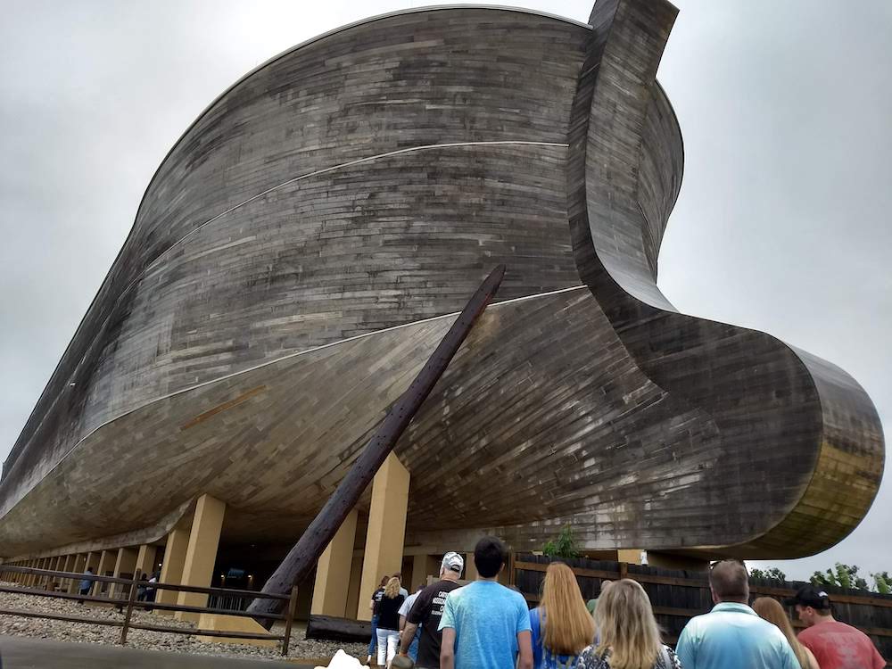 People are lined up outside. In front of them is a massive wooden boat resting on metal pillars.