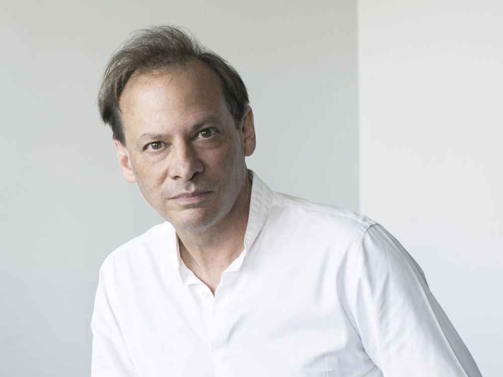 An older, light-skinned man wearing a white half-collared shirt against a white background looks into the camera.