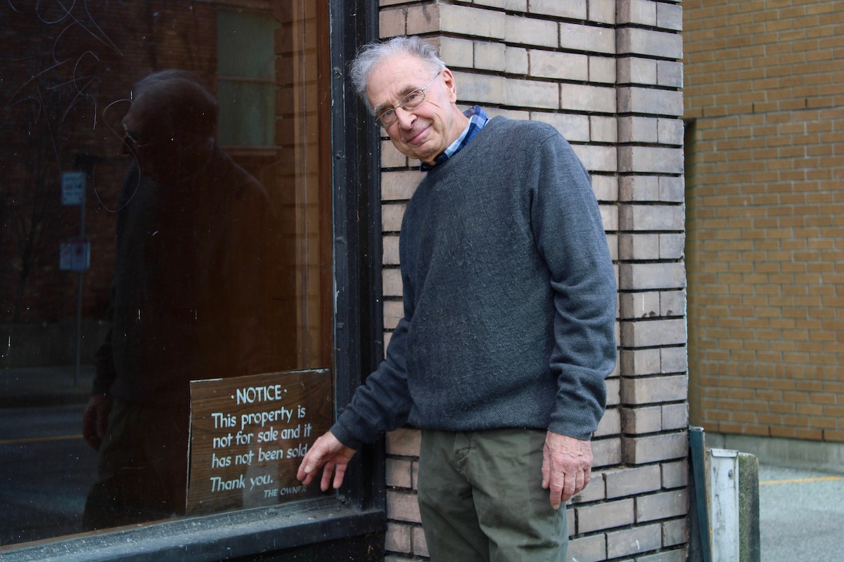 A man with light skin tone and grey hair wears a collared blue shirt and grey sweater. He smiles slightly at the camera as he lightly places his knuckles on a sign reading, 'NOTICE: This property is not for sale and has not been sold. Thank you. The owner.'