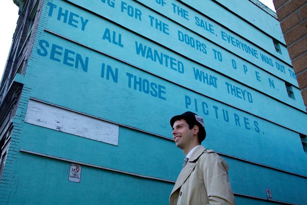 A young man with light skin, wearing a black hat, stands in front of the side of the Del Mar, which has been painted turquoise. The bottom two lines of the mural read, 'They All Wanted What They&rsquo;d Seen in Those Pictures.'