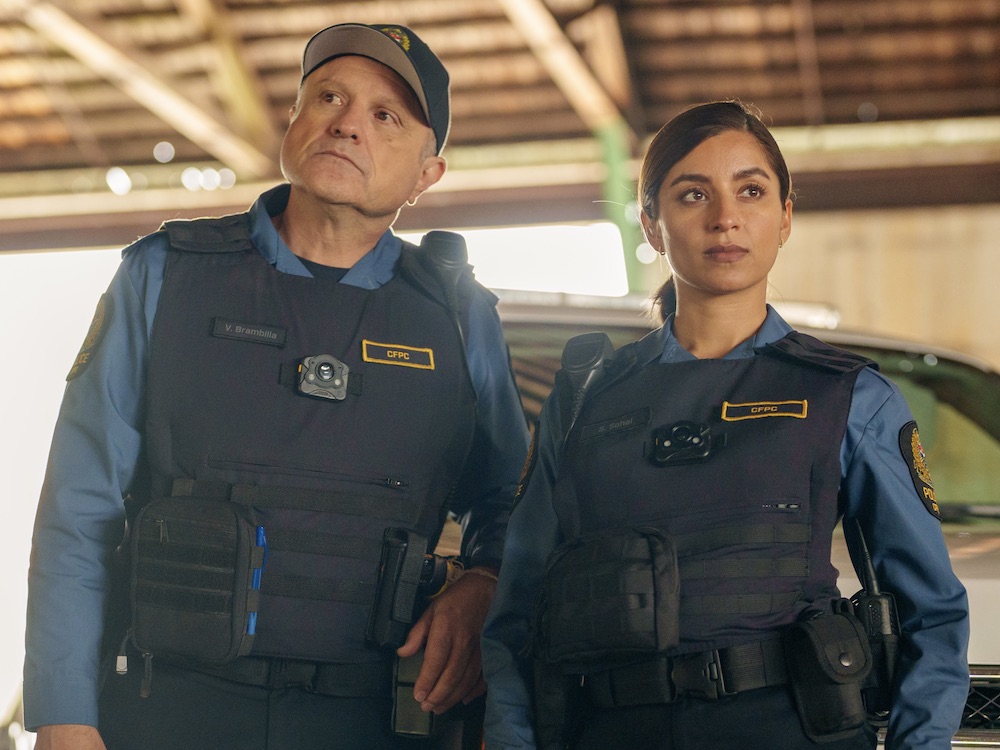 A television still features two people in dark blue police uniforms in a bright enclosed space. On the left, Enrico Colantoni is a taller man with light skin and brown eyes. On the right, Supinder Wraich is a South Asian woman with dark hair tied back and medium skin.