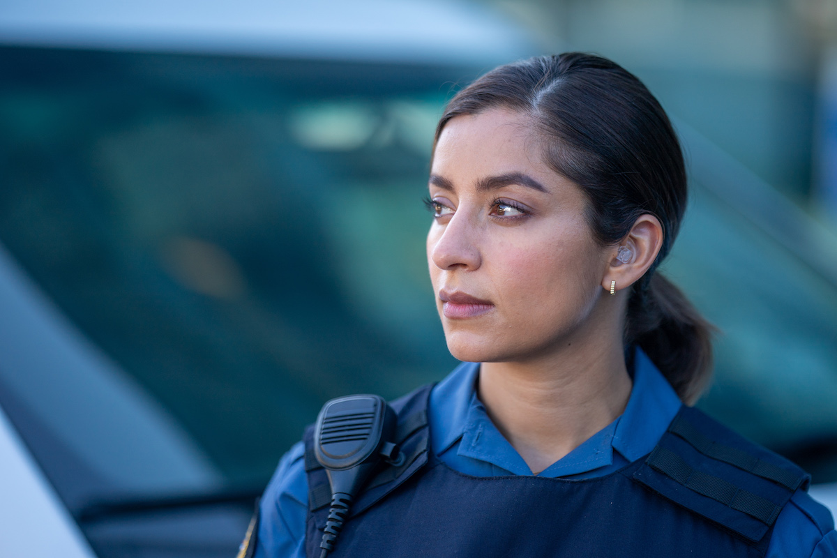 A television still of Supinder Wraich, a South Asian woman with her dark hair tied back, in a blue police uniform. She is standing in front of a police car in soft focus in the background, looking to the left of the frame.