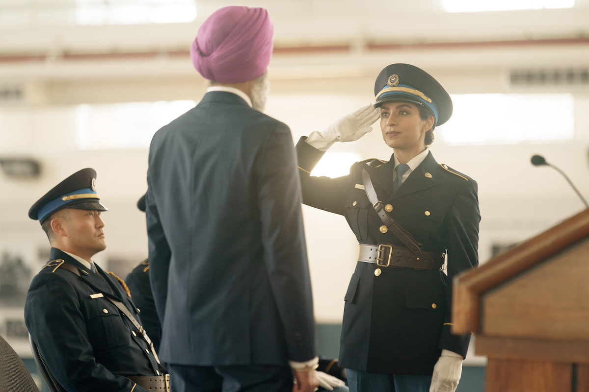 A television still features Supinder Wraich in a police uniform saluting a man in a turban with his back turned to the camera during a graduation ceremony.