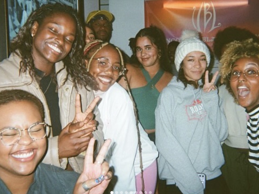 A group of young people with medium and dark skin are in a party environment, leaning towards each other and posing for the camera. In the background is a pink poster inscribed with the acronym 'VBL' for 'Vancouver Black Library.'