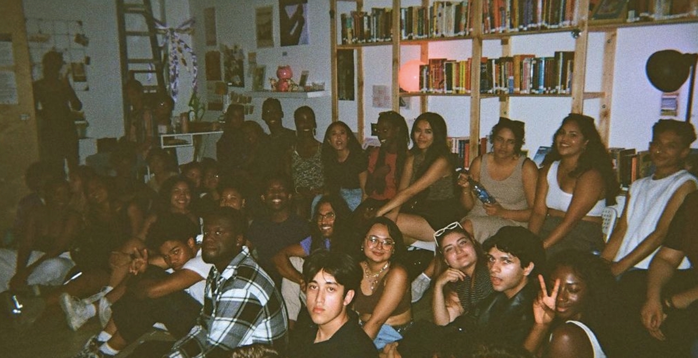 A crowd of young people are seated against a wall boasting a large bookshelf in an indoor performance venue. Many are looking at the camera.