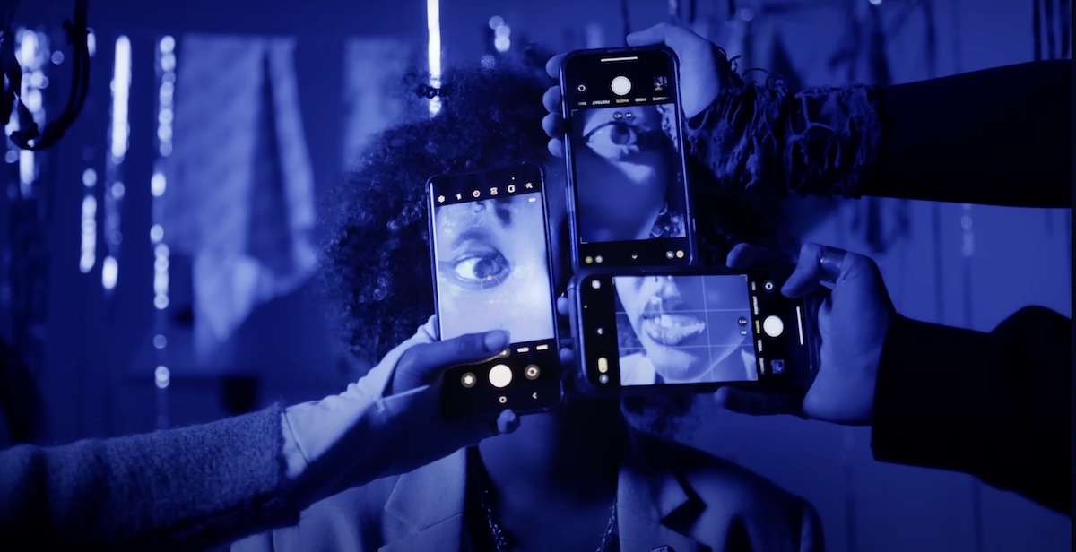 A monotone still, tinted dark blue, from a music video. Three hands hold phone cameras in front of a woman’s face. She has medium skin tone and curly hair.