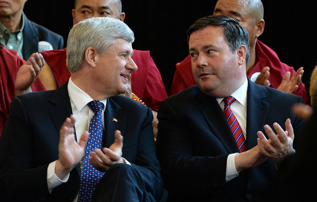 A grey-haired white man in a suit leans towards the dark-haired white man in a suit seated next to him at a large gathering, seemingly in conversation.