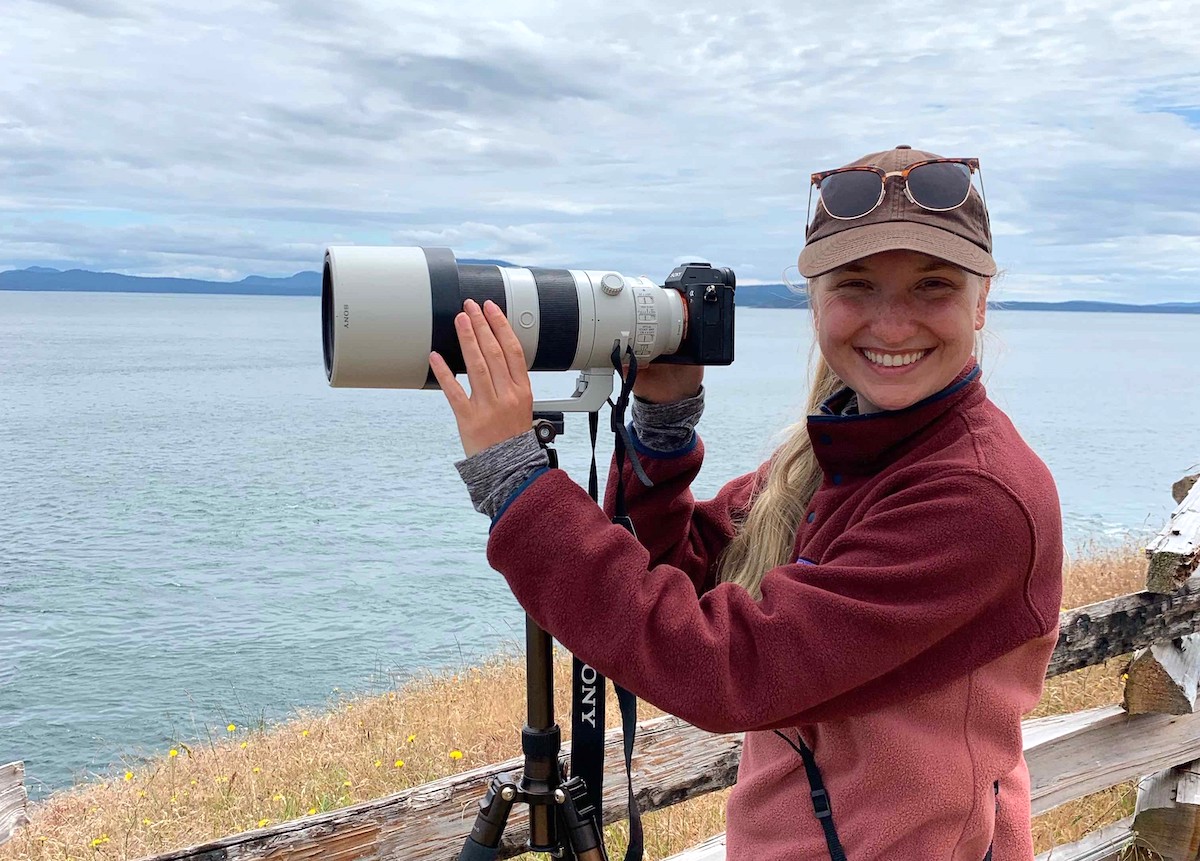 A woman with light skin tone and long blond hair, wearing a ball cap and fleece and touching a camera with a large lens on a tripod, smiles at the camera.