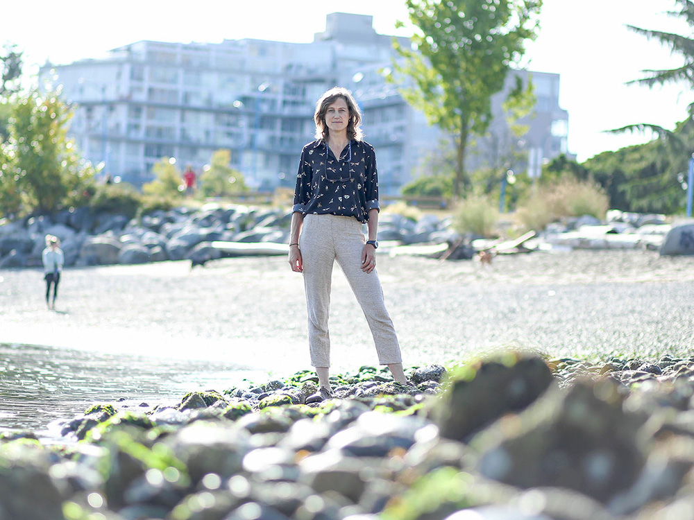 A woman with a light skin tone and shoulder-length brown hair stands on a rocky beach.