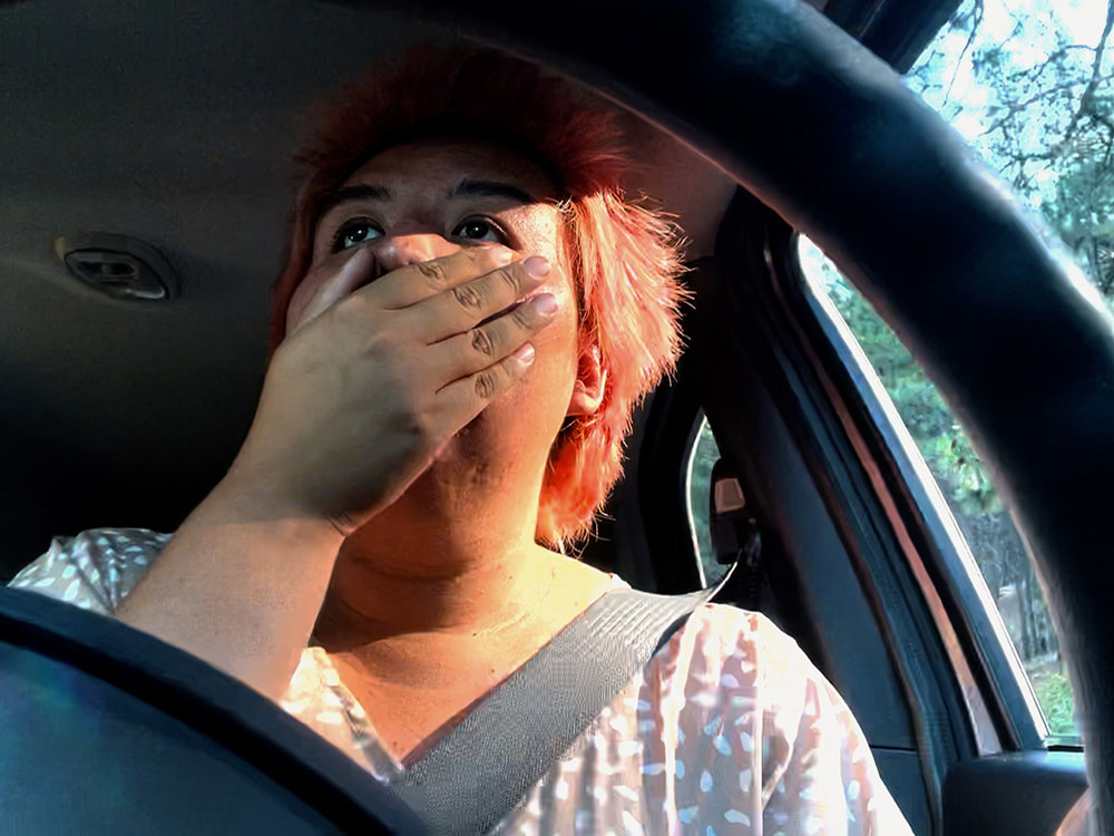 A woman with short dyed orange hair and medium skin tone claps her hand over her mouth. She is sitting in the driver’s seat of a car with the steering wheel in the foreground. She is looking at something up in the distance. Sun shines on the right side of her face.