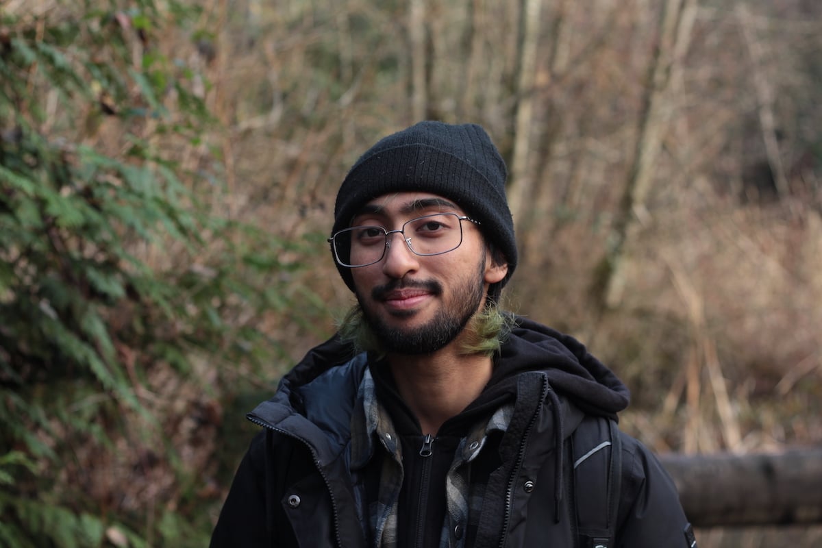A man with medium skin tone in his 20s wears a dark hoodie, a blue flannel shirt and jacket. He has glasses, a beard and tufts of green hair sticking out of his dark toque. He is surrounded by the green and brown hues of a forest around him.
