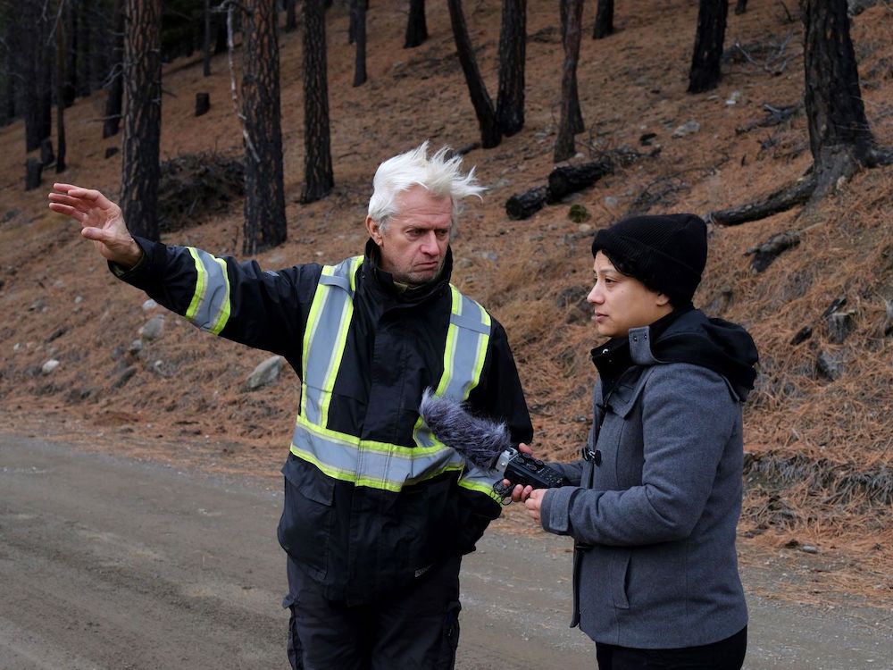 On the right, a woman in a black toque and grey jacket, holding a recorder with a wind muffed microphone in her hand, looks off to the left of the frame, to where the man standing beside her is pointing. He has white hair blowing in the wind and wears a black jacket with high-vis striping. The charred trunks of trees can be seen behind them.