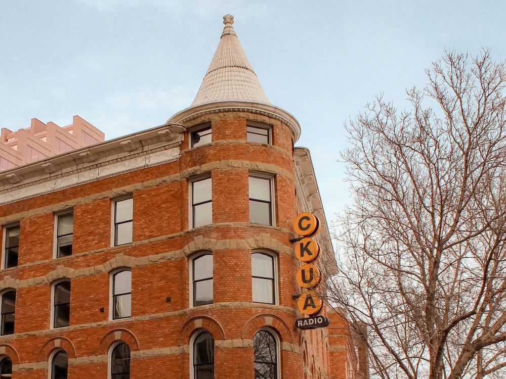 An exterior shot of a red brick building with a small turret. The station&rsquo;s call sign, CKUA, appears in yellow circles on a sign attached to the corner of the building.