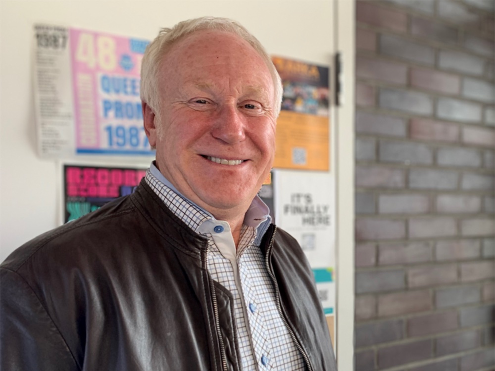An older man with short white-blond hair and a light skin tone smiles at the camera. He is standing in a Calgary caf&eacute;.