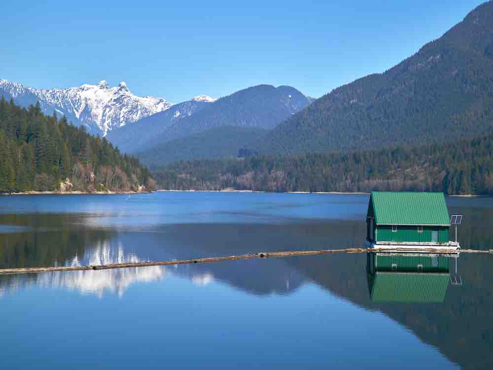 A serene lake features a small green hut in the middle of the water. Dark blue snow-capped mountains rise in the background against a bright blue sky on a sunny day.