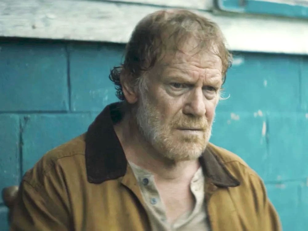 A man in his 60s with thin dirty-blond hair and light skin stands against a teal brick wall. He wears a brown work jacket over a beige Henley and looks grimly into the distance.