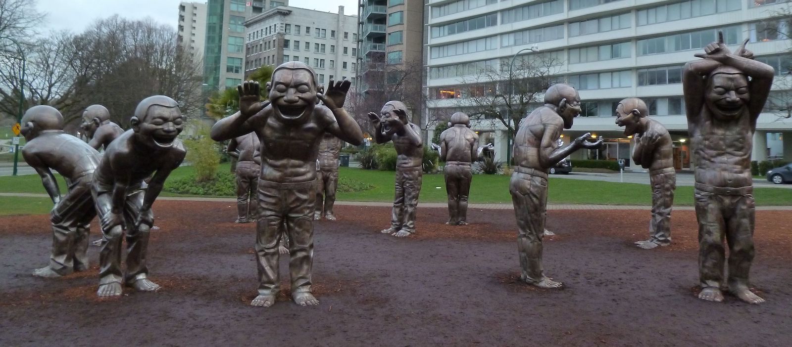 A series of bronze sculptures of short-haired, shirtless men in different poses of laughter. One in the foreground holds his thumbs to his ears, palms open. Others are bent over laughing.