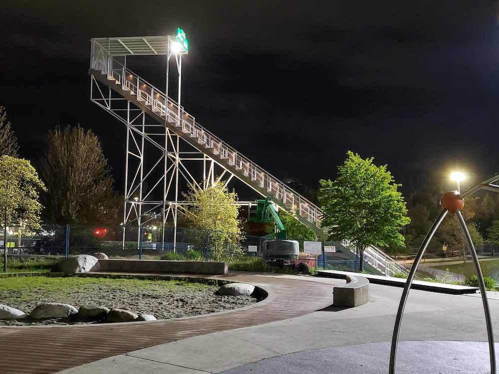 A large white set of bleachers stands alone in a public park against a night sky with a wooden walkway in the foreground.