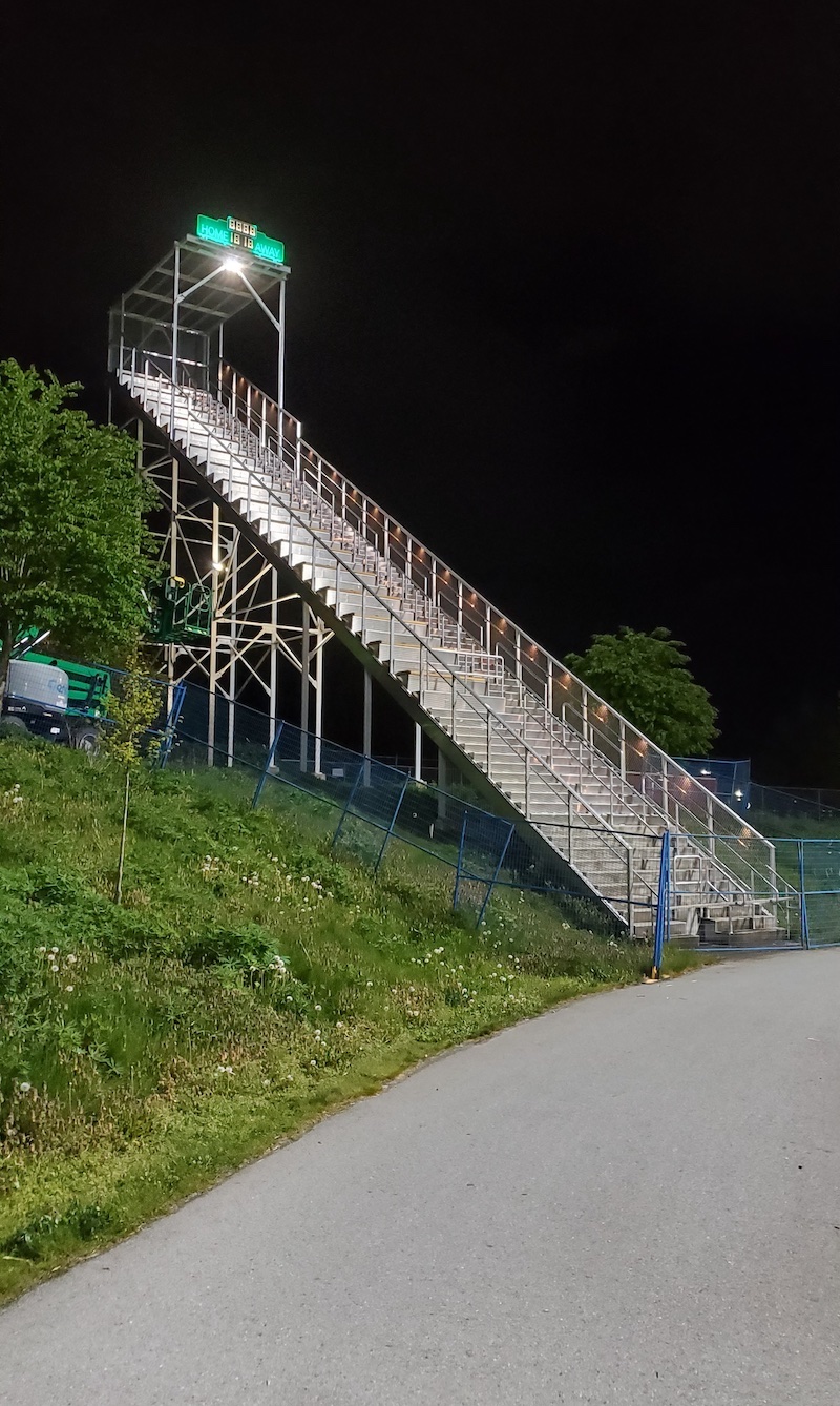 A tall, narrow grandstand with a green awning and light at the top descends down a gentle grassy slope against a night sky.