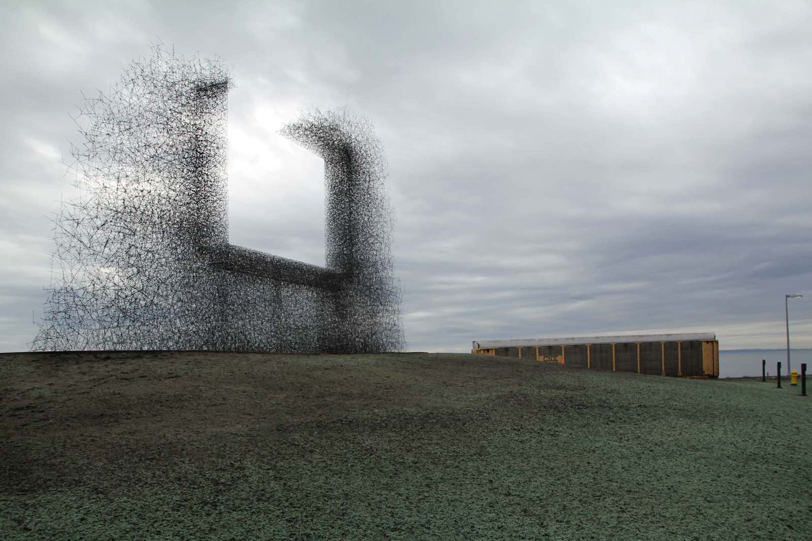 A large public art installation composed of dark wiry materials creates the shape of a staticky metal picture frame without a top. It stands against a grey sky.