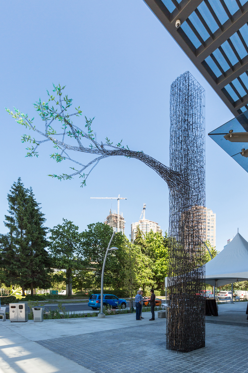 A large, wiry public art installation in the shape of a tree with one branch features small, bright green leaves.