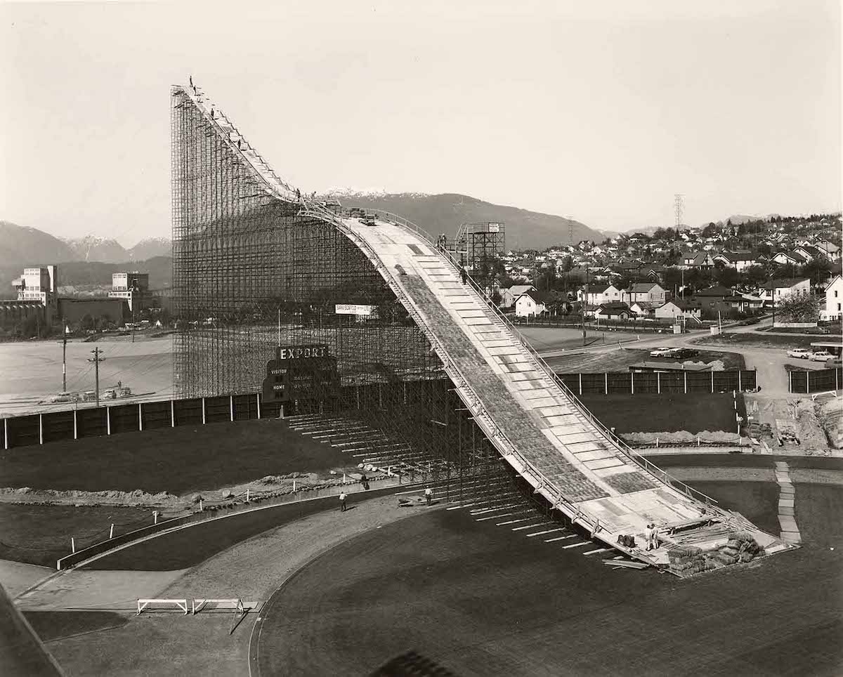 A black and white archival photo depicts a giant wooden ski jump over a sports field with grass and a running track around it.
