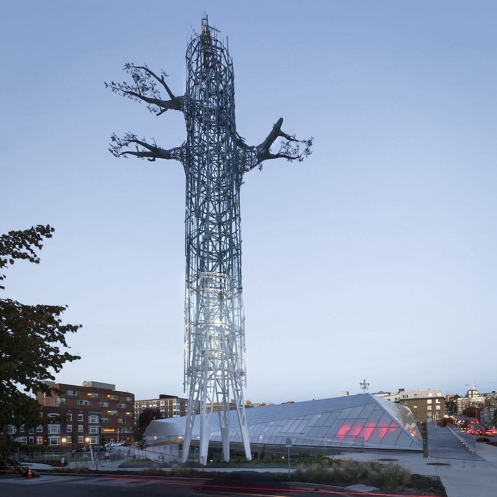 A tall piece of public art depicts a metallic tree with three small branches near its top against a blue sky at twilight.