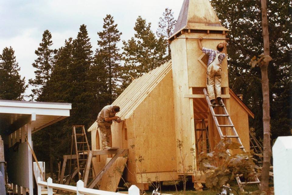 An aging photo with yellow hues shows two men constructing a small building with plywood. There are conifer trees behind.