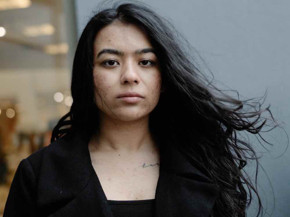 A young woman with long dark hair, dark eyes and light skin looks solemnly towards the camera. She is wearing black and standing against a glass wall in soft focus.