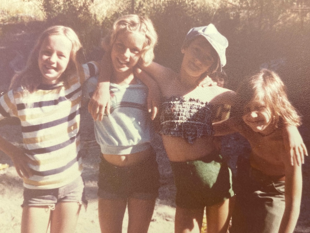 Four school-aged children pose in golden summer light on a film photograph from the 1970s. They are wearing summer clothes and have their arms around each other.